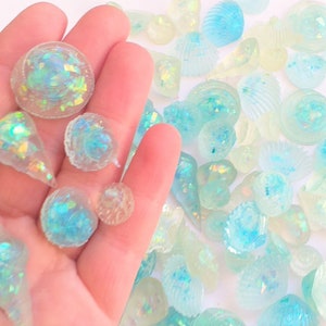 May include: A collection of iridescent blue and white seashells, some with a pearlescent finish, are scattered on a white surface. A hand is holding a few of the shells.