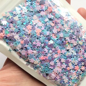 May include: A bag of iridescent glitter featuring a mix of shapes, including stars, hearts, and flowers. The glitter is in shades of blue, purple, and pink.