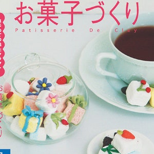 May include: A book cover with a colorful illustration of various miniature pastries made from clay. The title of the book is "O-Kashi-Tsukkuri" which translates to "Making Sweets" in English. The book cover also includes the text "Patisserie De Clay" and "300 Yen".