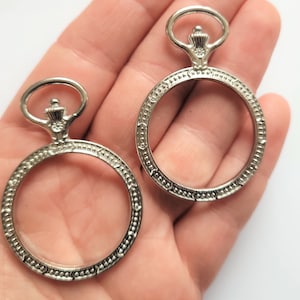 May include: Two silver-toned metal watch face blanks with ornate bezels and a loop at the top for hanging.