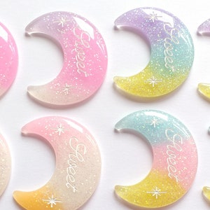 May include: Six pastel-coloured crescent moon shaped resin ornaments with glitter and the word "Sweet" written on each one.