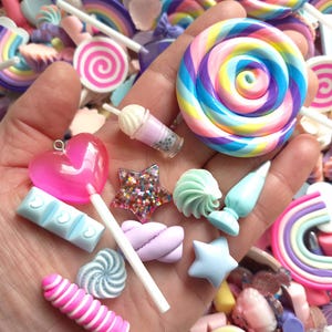 Può includere: Una mano mostra una varietà di caramelle e dolci in miniatura colorati. Sono inclusi un grande lecca-lecca a spirale, un lecca-lecca a forma di cuore rosa, una stella con glitter e una mini bevanda. Altre caramelle sono sparse sullo sfondo.