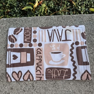 May include: A white cotton kitchen towel with a brown and pink coffee themed design. The towel features the words "Java", "Mocha", "Cappuccino", and "Latte" in a stylized font.