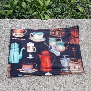 May include: A rectangular, black fabric placemat with a coffee theme. The placemat features watercolor illustrations of coffee cups, teapots, a coffee grinder, and a bag of coffee beans. The illustrations are in shades of red, blue, brown, and white.