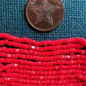 May include: A close-up of a strand of bright red square beads. The beads are small and uniform in size. A copper-colored coin with a starfish design and the words "ONE CENT" is visible in the background.