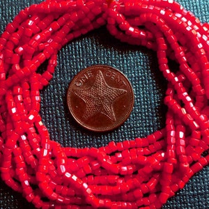 May include: A close-up of a red square bead necklace. The necklace is made of many small, square beads that are strung together. The beads are a bright, vibrant red color. The necklace is lying on a dark blue surface. The necklace is in the shape of a circle, and there is a one cent coin in the center of the circle.
