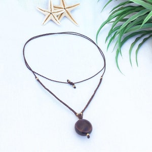 May include: A brown macrame necklace with a circular pendant. The necklace features a sliding knot closure and small gold-colored beads. Two starfish and green foliage are in the background.