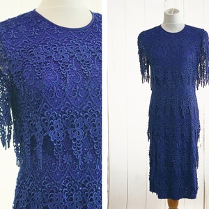 May include: A blue lace dress with a tiered skirt and fringe detailing. The dress has a round neckline and short sleeves.