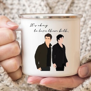 May include: White enamel mug with a black and white illustration of two men and the text "It's okay to love them both."