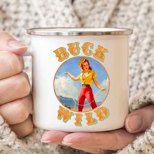 May include: White enamel mug with a colorful illustration of a woman in a yellow shirt and red pants holding a lasso. The text "BUCK WILD" is printed in orange and blue letters around the illustration.
