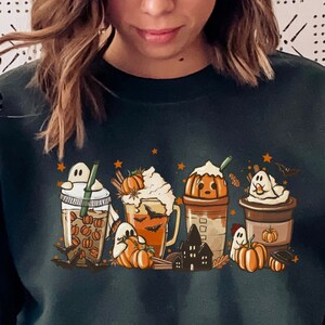 Könnte beinhalten: Ein dunkelgrüner Pullover mit einem Aufdruck von fünf Halloween-Kaffeegetränken. Die Getränke sind mit Kürbissen, Geistern und Sternen verziert.