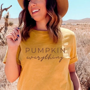 Puede incluir: Una mujer lleva una camiseta amarilla mostaza con el texto "Pumpkin everything" impreso en ella. Lleva un sombrero marr&oacute;n y unos vaqueros azules. La mujer est&aacute; de pie en un paisaje des&eacute;rtico.