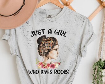 Può includere: Una maglietta grigia con una grafica di una donna con libri impilati sulla testa. Il testo "Just a girl who loves books" è stampato sotto la grafica.