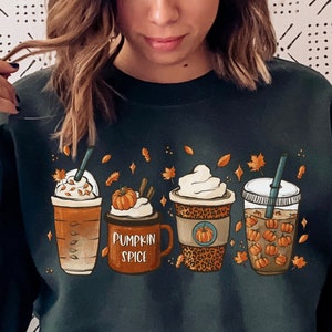 May include: A dark green sweatshirt with a graphic of four fall-themed drinks. The drinks are a caramel latte, a pumpkin spice latte, a coffee with whipped cream, and a cold brew with pumpkin spice. The text "Pumpkin Spice" is printed on the sweatshirt.