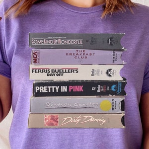 Puede incluir: Camiseta morada con una pila de cintas VHS con los títulos "Some Kind of Wonderful", "The Breakfast Club", "Ferris Bueller's Day Off", "Pretty in Pink", "Sixteen Candles" y "Dirty Dancing".
