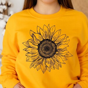 Könnte beinhalten: Ein gelber Pullover mit einem schwarzen Sonnenblumen-Grafikdruck. Die Sonnenblume ist mit einer schwarzen Umrandung und einer schwarzen Mitte detailliert.