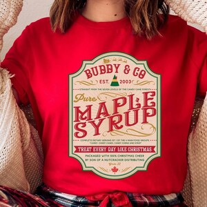 May include: Red t-shirt featuring a vintage-style label that reads "Bubby & Co. Est. 2003 Pure Maple Syrup" with a green elf hat graphic. The label has a green border and text that reads "Straight from the Seven Levels of the Candy Cane Forest. Complete Dietary Serving of 1 of the 4 Main Food Groups. Treat Every Day Like Christmas. Packaged with 100% Christmas Cheer by Son of a Nutcracker Distributing."