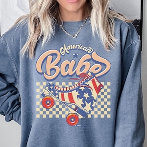 May include: A blue sweatshirt with the text "American Babe" and a graphic of a roller skate in red, white, and blue. The roller skate design includes stars and stripes, set against a checkered background. The brand name "Blue Jern" is in the top left corner.