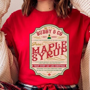 May include: Red t-shirt featuring a vintage-style label that reads "Bubby & Co. Est. 2003 Pure Maple Syrup" with a green elf illustration. The label has text that reads "Straight from the Seven Levels of the Candy Cane Forest. Complete Dietary Serving of 1 of the 4 Main Food Groups. Treat Every Day Like Christmas. Packaged with 100% Christmas Cheer by Son of a Nutcracker Distributing."