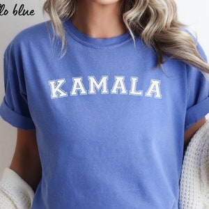 May include: A flo blue t-shirt with the name "KAMALA" printed in white block letters. The shirt has short sleeves and a crew neck. The text "flo blue" is in the upper left corner.