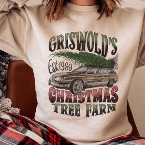 May include: A beige sweatshirt with a vintage-style graphic print of a station wagon with a Christmas tree on top. The text reads "Griswold's Est. 1989 Christmas Tree Farm".