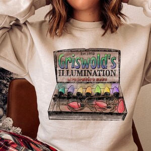 May include: A sand-colored sweatshirt featuring a graphic of a vintage Christmas light box with the text "Griswold's Illumination Decorative Sets" and the year "Est. 1989". The box contains colorful light bulbs.