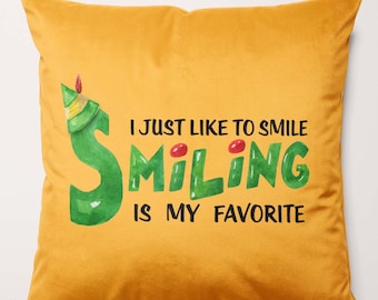 Puede incluir: Una almohada de tiro dorada con una ilustración verde y roja de un elfo sonriente. El texto en la almohada dice "I just like to smile smiling is my favorite".