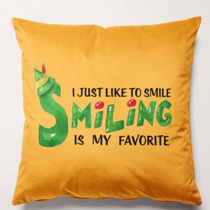 Op de afbeelding: Een gouden kussen met een groene en rode illustratie van een lachende elf. De tekst op het kussen luidt "I just like to smile smiling is my favorite".