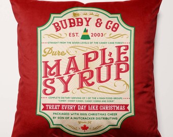 Puede incluir: Cojín decorativo rojo con un diseño de etiqueta de estilo vintage. La etiqueta dice "Bubby & Co. Pure Maple Syrup" con el texto "Treat Every Day Like Christmas". Un artículo de decoración del hogar festivo.