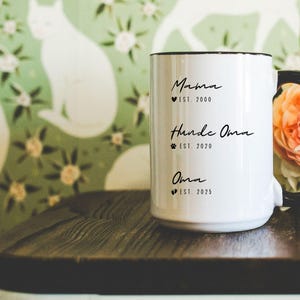 Könnte beinhalten: Weiße Keramiktasse mit schwarzem Rand und schwarzem Henkel. Die Tasse trägt den Text "Mama est. 2000", "Handke Oma est. 2020" und "Oma est. 2025".