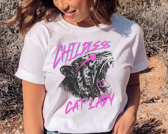 May include: White t-shirt with a black and white graphic of a roaring panther. The graphic is surrounded by pink and blue text that reads "Childless Cat Lady".