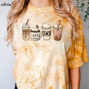 May include: A yellow tie-dye t-shirt with a graphic of four different coffee drinks. The text "Cat Mom" is on one of the coffee cups.