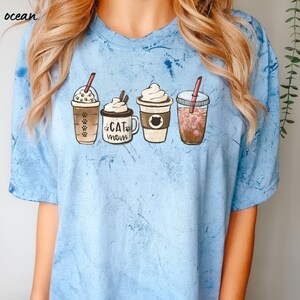 May include: A light blue t-shirt with a tie-dye pattern featuring four illustrated coffee cups. The cups have different designs, including a cat, paw prints, and hearts. The text "Cat Mom" is on one of the cups.