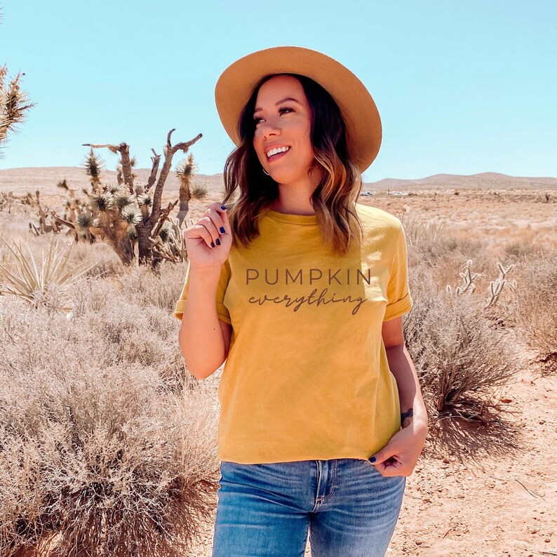 Puede incluir: Una mujer lleva una camiseta amarilla mostaza con el texto "Pumpkin everything" impreso en ella. Lleva un sombrero marr&oacute;n y unos vaqueros azules. La mujer est&aacute; de pie en un paisaje des&eacute;rtico.