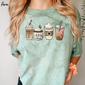 May include: A light green t-shirt with a graphic of four different coffee drinks. The drinks are a iced coffee, a hot chocolate, a latte, and a pink drink. The text "Cat Mom" is printed on the hot chocolate.