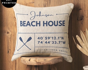 May include: A beige square cushion with a navy blue border and text. The text reads "Johnson Beach House", "40°59'12.1"N 74°44'33.7"W", and "Established 2013". There is a graphic of two crossed oars in the centre of the cushion.
