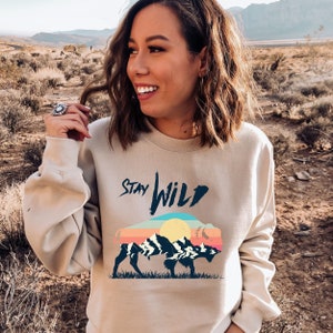 Könnte beinhalten: Ein beiger Pullover mit dem Aufdruck "Stay Wild" auf der Vorderseite. Der Text ist in schwarzer, handschriftlicher Schrift. Unter dem Text befindet sich eine Grafik eines Bisons mit einem Sonnenuntergang und Bergen im Hintergrund. Der Bison ist schwarz und der Sonnenuntergang ist ein Farbverlauf von Rosa, Orange und Gelb.