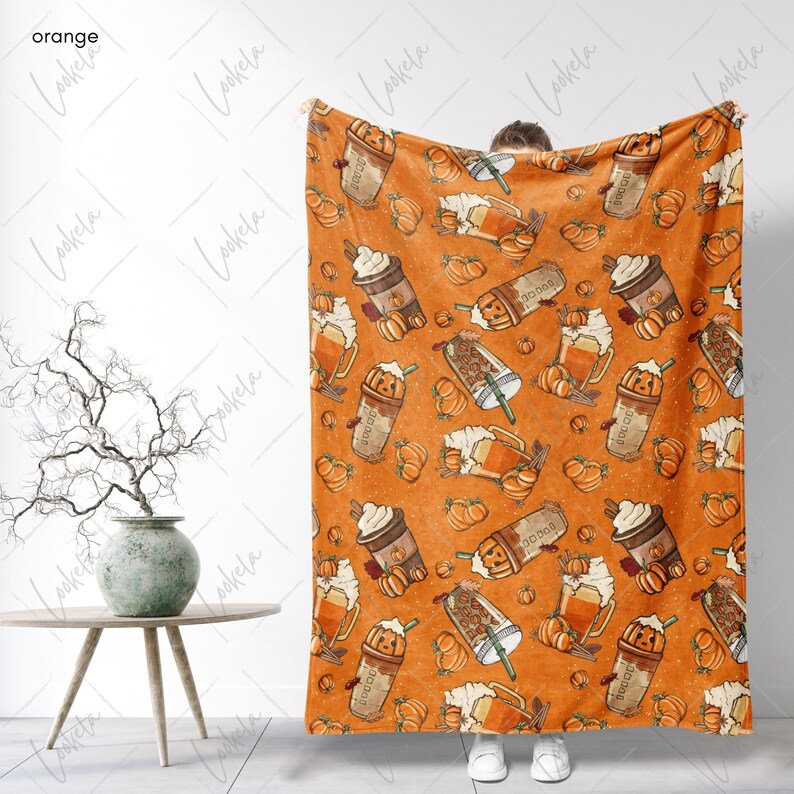 Op de afbeelding: Een oranje fleece deken met een patroon van pompoenen, koffiebekers en mokken. Het patroon wordt over de hele deken herhaald.