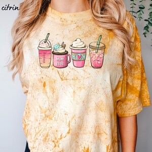 Puede incluir: Una camiseta amarilla tie-dye con un gr&aacute;fico de cuatro bebidas de caf&eacute; diferentes. Las bebidas est&aacute;n etiquetadas como "Mom Fuel", "Mom" y tienen corazones en ellas.