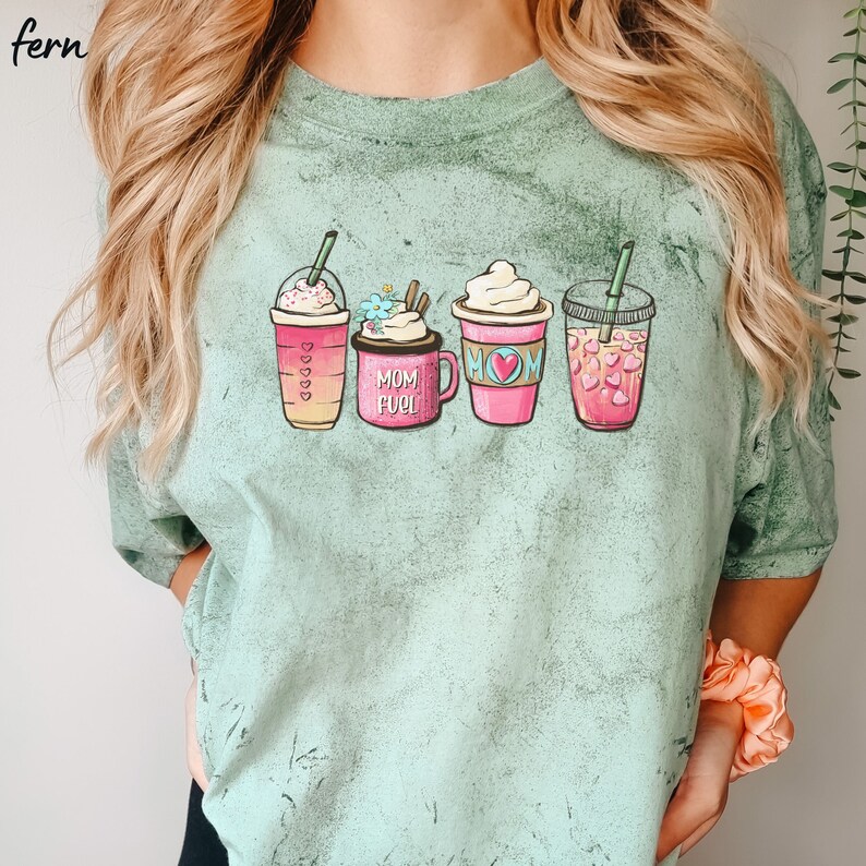 Puede incluir: Una camiseta verde claro con un gr&aacute;fico de cuatro bebidas de caf&eacute; diferentes. Las bebidas est&aacute;n etiquetadas como "Mom Fuel" y tienen corazones y flores.