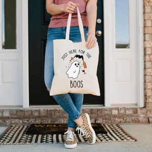 Könnte beinhalten: Eine weiße Canvas-Tragetasche mit einer schwarzen und orangefarbenen Grafik eines Geistes, der eine Weinflasche und ein Glas hält. Der Text auf der Tasche lautet "Just here for the boos".