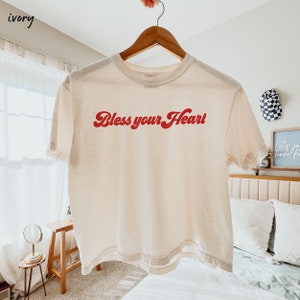 Op de afbeelding: Een gebroken wit getailleerd T-shirt met de tekst "Bless your Heart" in rode letters.