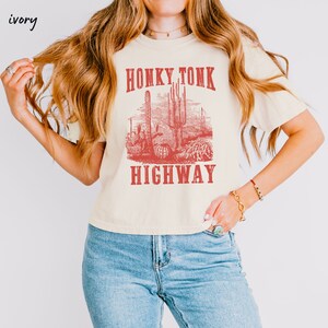May include: A cream colored t-shirt with a red graphic print of a desert scene with cacti and the text "Honky Tonk Highway".