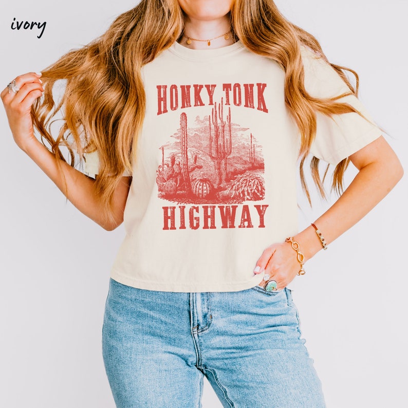 May include: A cream colored t-shirt with a red graphic print of a desert scene with cacti and the text "Honky Tonk Highway".