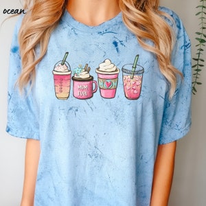 Puede incluir: Una camiseta azul claro con un gr&aacute;fico de cuatro bebidas de caf&eacute; diferentes. Las bebidas est&aacute;n todas en vasos rosas con tapas blancas y pajitas. Uno de los vasos tiene el texto "MOM FUEL".