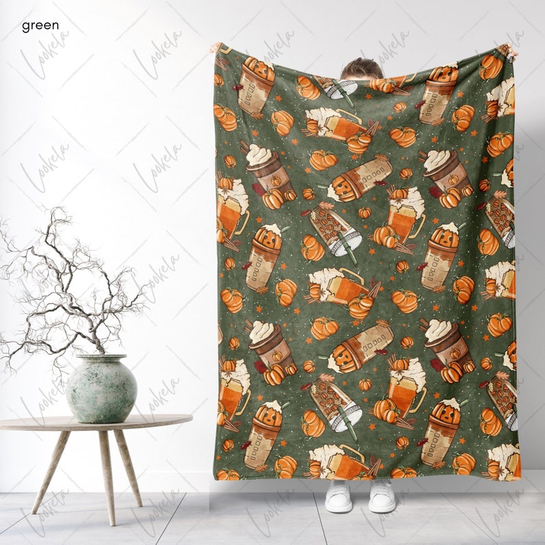 Op de afbeelding: Een groene fleece deken met een patroon van oranje pompoenen, bruine koffiebekers en witte sterren. De deken wordt vastgehouden door een persoon, waardoor het volledige patroon zichtbaar is.