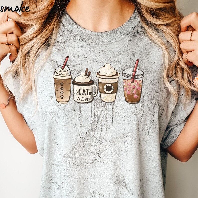 May include: A light gray t-shirt with a graphic of four coffee drinks. The drinks are a iced coffee, a latte, a cappuccino, and a boba tea. The text "Cat Mom" is printed on the shirt.