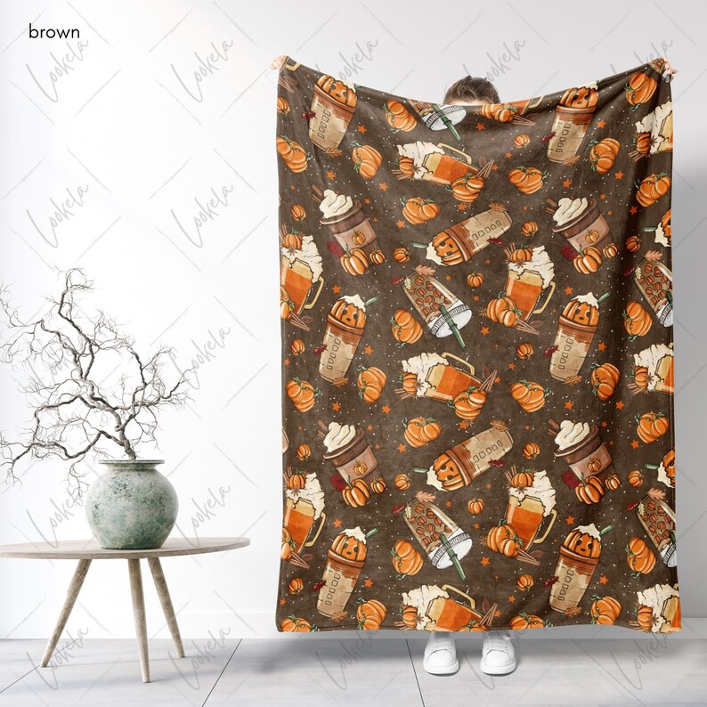 Op de afbeelding: Een bruine fleece deken met een patroon van pompoenen, koffiebekers en sterren. De koffiebekers hebben een pompoen gezicht en zeggen "Pumpkin Spice" aan de zijkant.