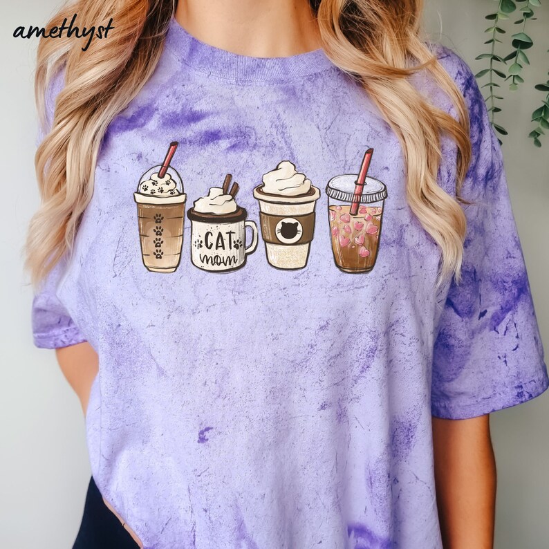 May include: Purple tie-dye t-shirt with a graphic of four coffee cups. The cups have different designs, including a cat, paw prints, and whipped cream. The text "Cat Mom" is on one of the cups.
