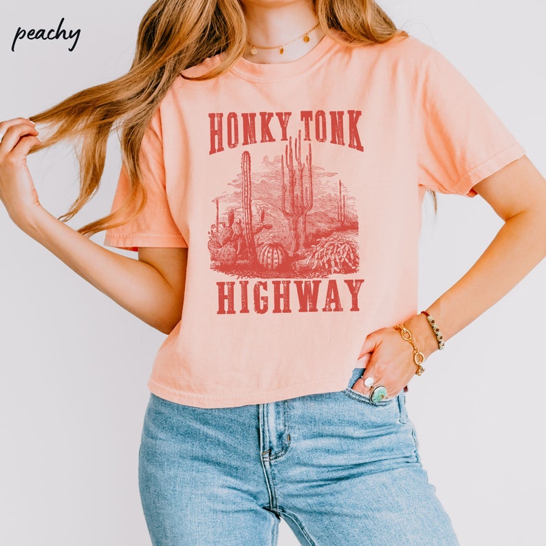 May include: Peach colored cropped t-shirt with a vintage style graphic of a desert scene with cacti and the text "Honky Tonk Highway" in red.